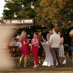 Ludzie na imprezie plenerowej przy grillu i piwie, rozmawiający przy stołach i białych obrusach.