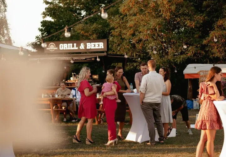 Ludzie na imprezie plenerowej przy grillu i piwie, rozmawiający przy stołach i białych obrusach.