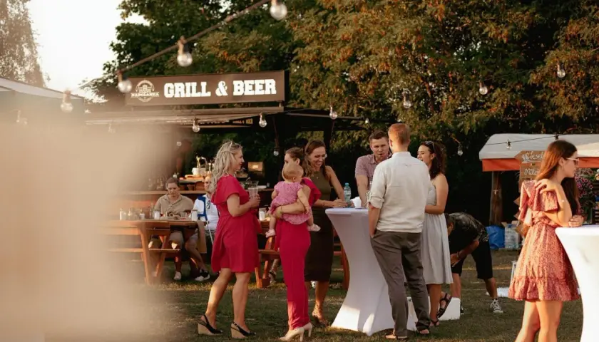Ludzie na imprezie plenerowej przy grillu i piwie, rozmawiający przy stołach i białych obrusach.