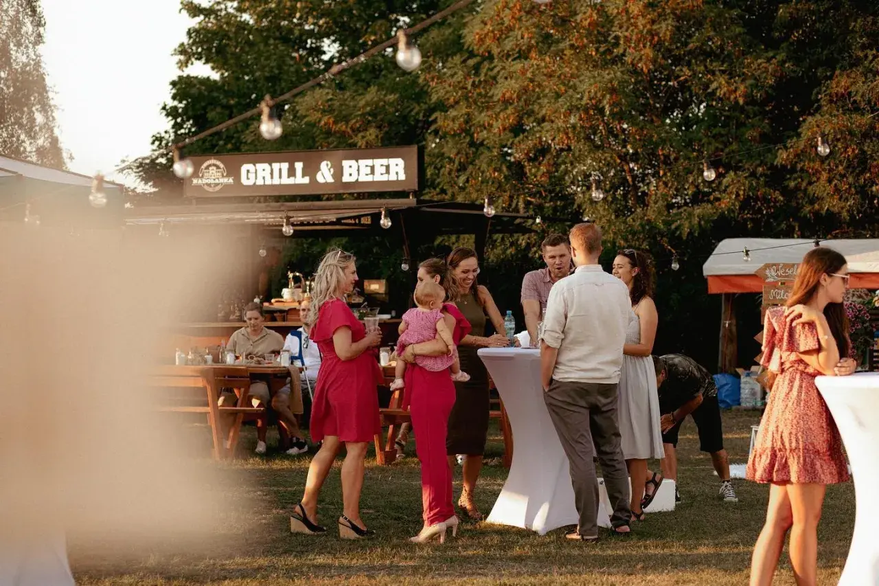 Ludzie na imprezie plenerowej przy grillu i piwie, rozmawiający przy stołach i białych obrusach.